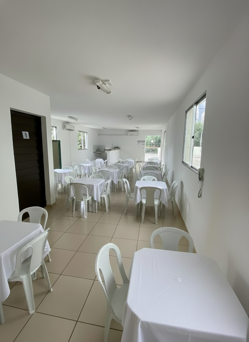 Salão de festas com mesas brancas com toalhas, cadeiras plásticas, piso em cerâmica e ar condicionado